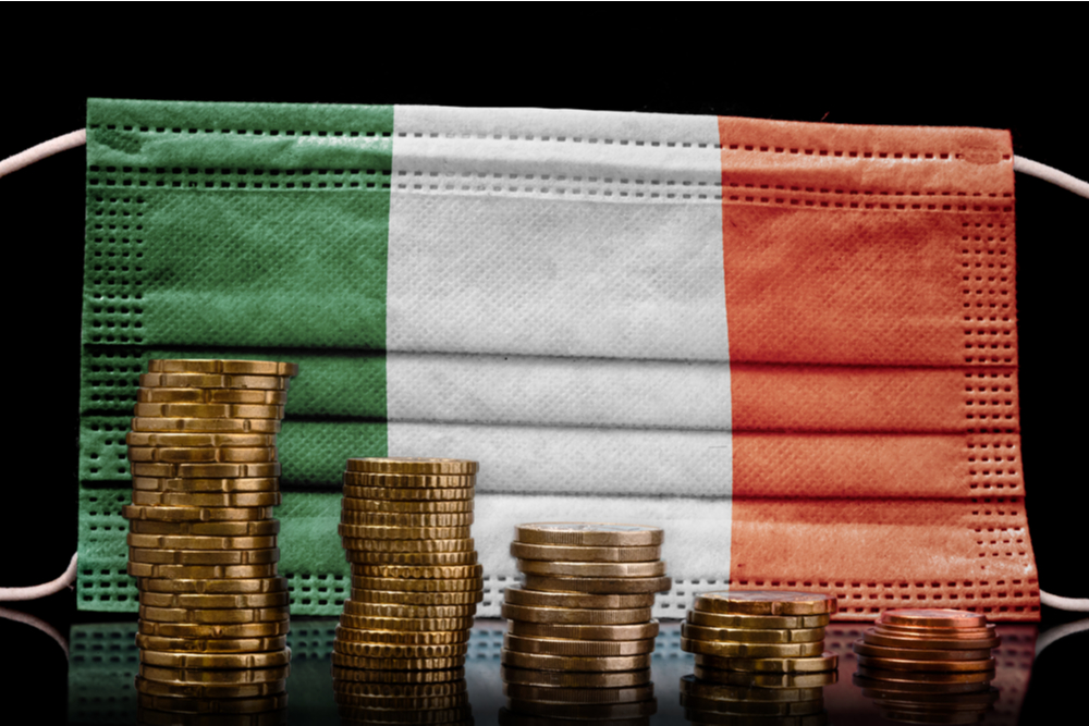 A surgical mask with the flag of Ireland behind some descending stacks of various coins.