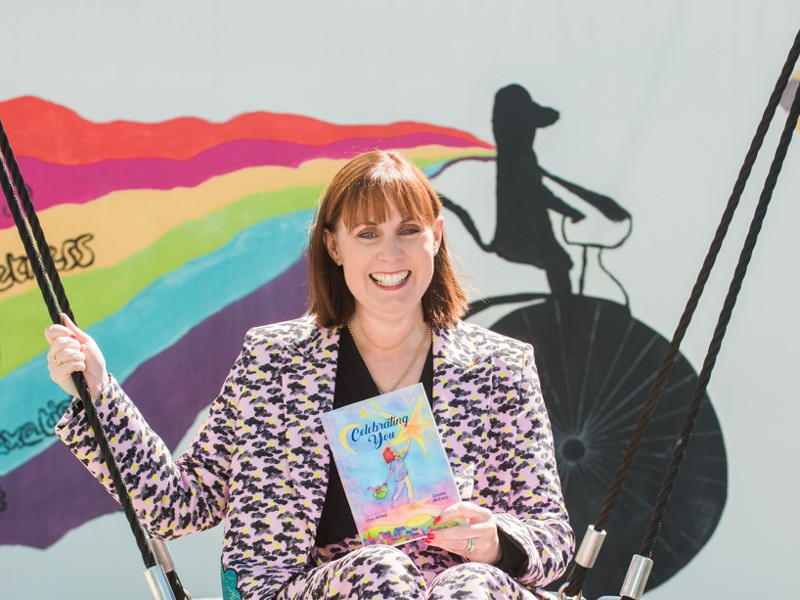 Woman in brightly coloured coat sitting on a swing holding a book.