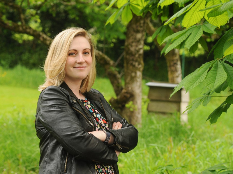 Woman with blonde hair standing beside a bee hive.