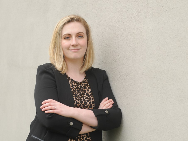 Woman in black jacket, with blonde hair, leaning against a wall.