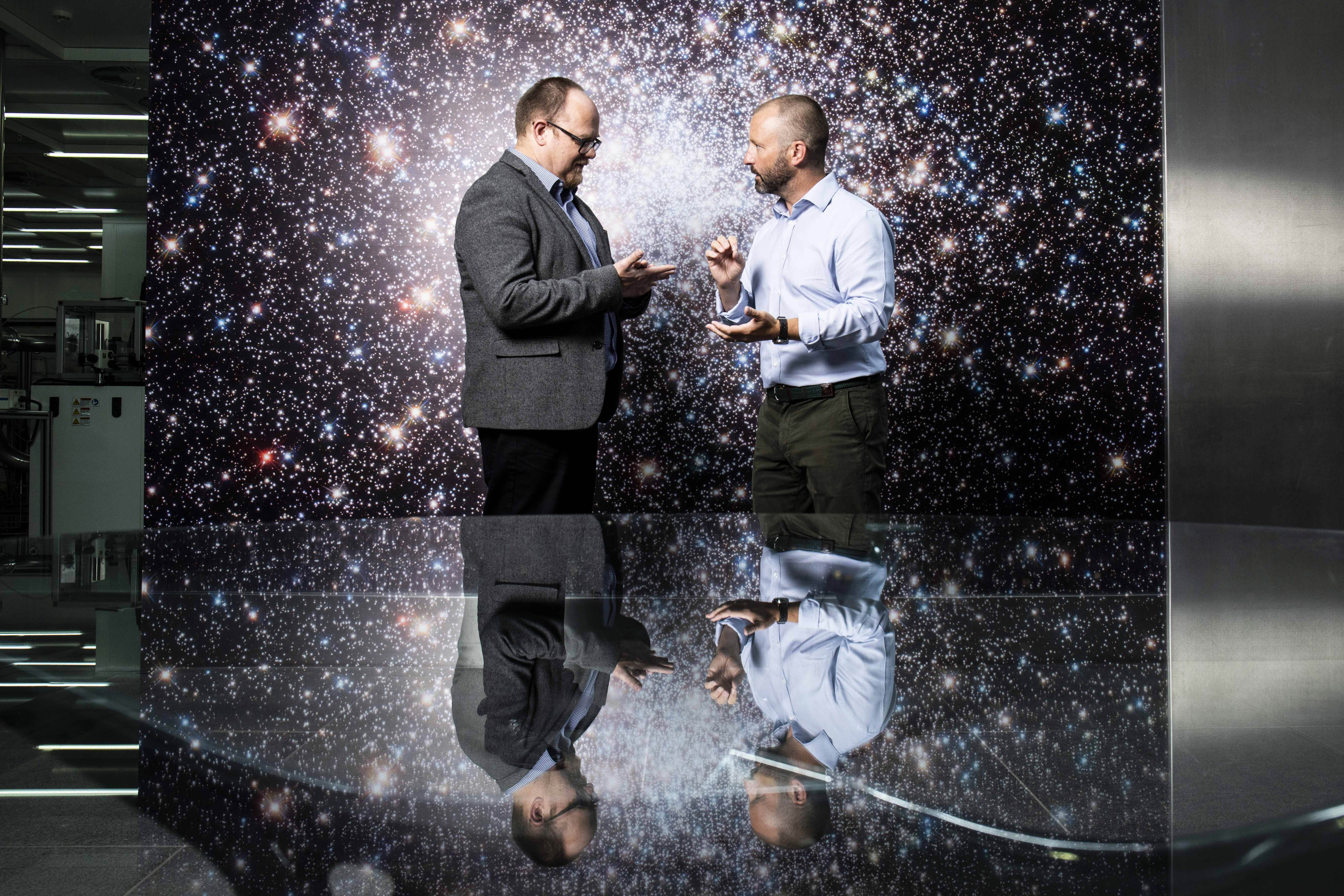 Two men stand talking in a boardroom surrounded by stars.