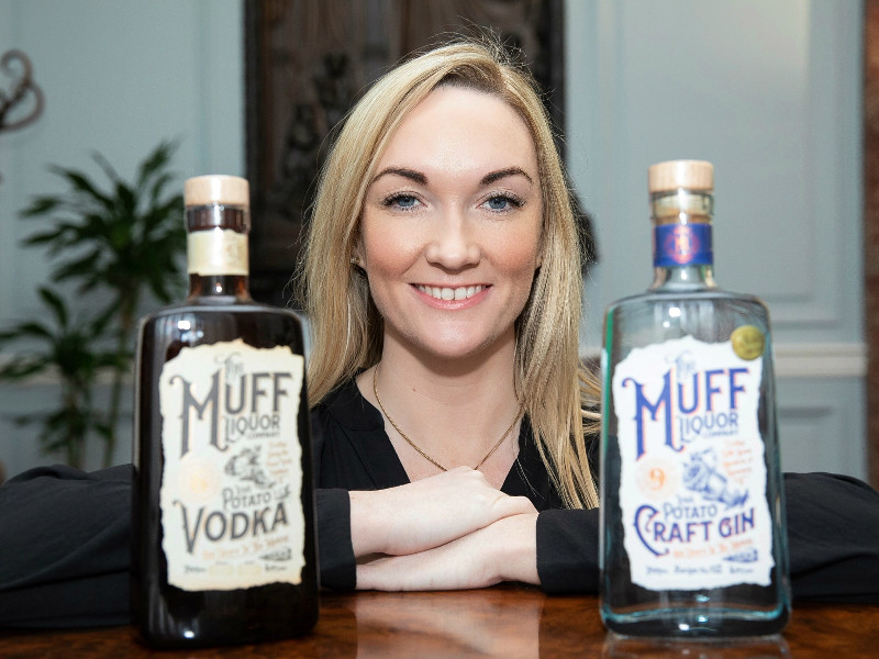 Blonde-haired woman stands in between a bottle of Irish gin and vodka.