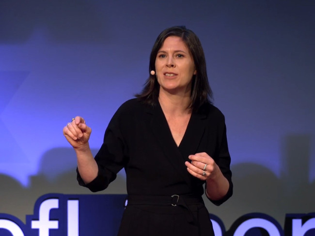 Woman in black dress talking on a stage at a TED talk.