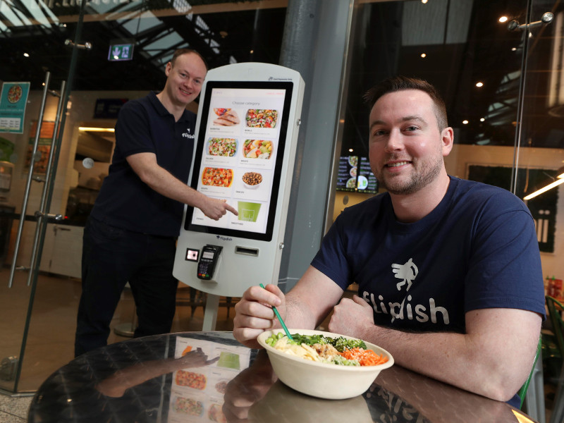 Man standing beside kiosk while man at foreground eats noodles.