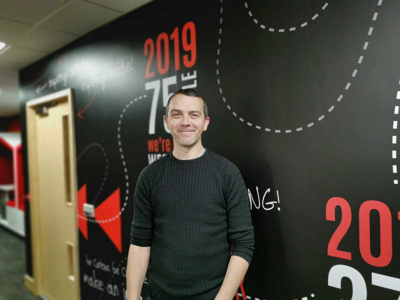 Man in black jumper stands in front of wall with murals.