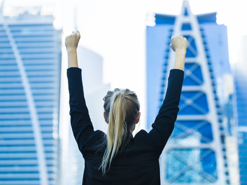 Blonde woman in business suit holds hands in air in jubilation.