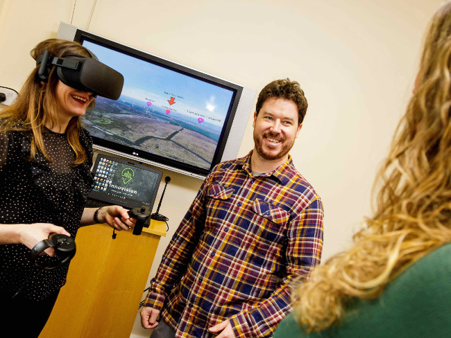 Man in check shirt standing beside woman wearing a VR headset.