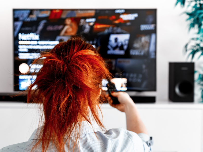 Red-haired woman using a remote control to watch Netflix on her smart TV.