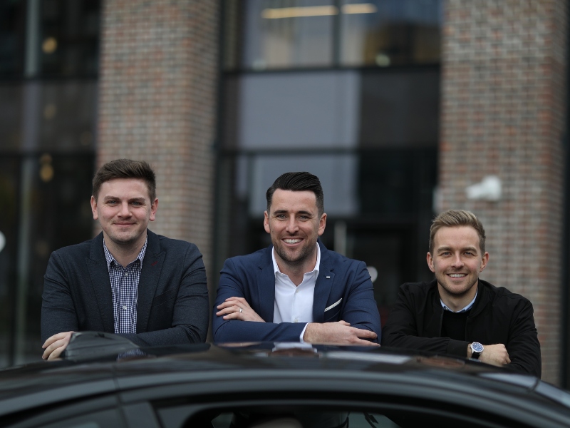 Three young men lean on top of a car.