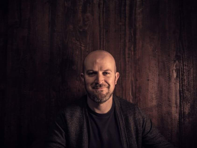 Man in jacket sitting in front of a wooden wall.