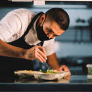 male chef with mask decorating food in ceramic dishes over stainless steel worktop in restaurant kitchen during coronavirus pandemic.
