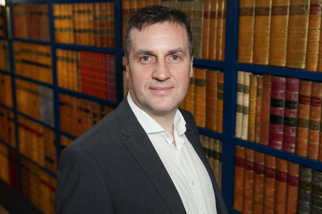 Man in dark suit, white shirt, standing in library.