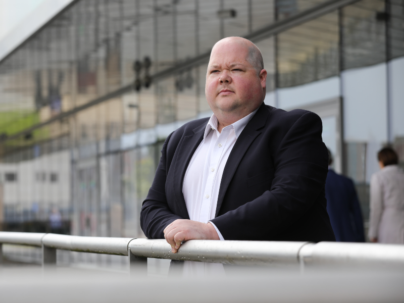 Man in black jacket, white shirt, leaning against a rail.