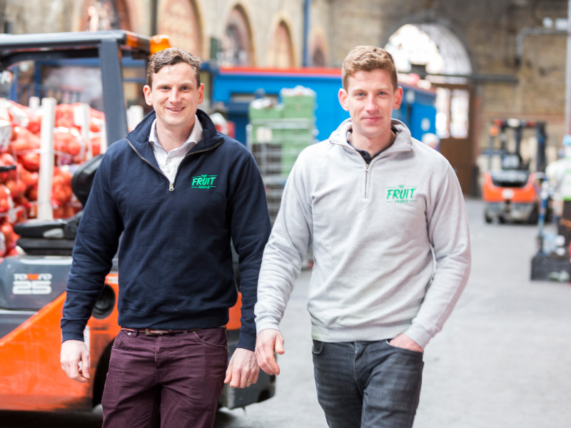 Two young men walking through a fruit market.