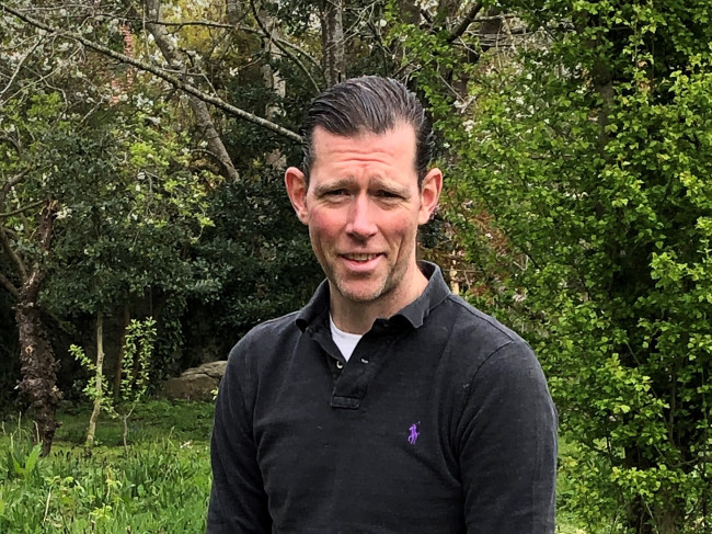 Dark-haired man standing beside trees.