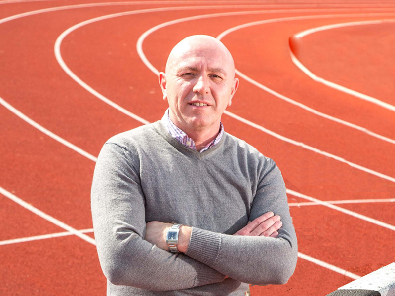 Man in grey jumper, arms folded, on a running track.