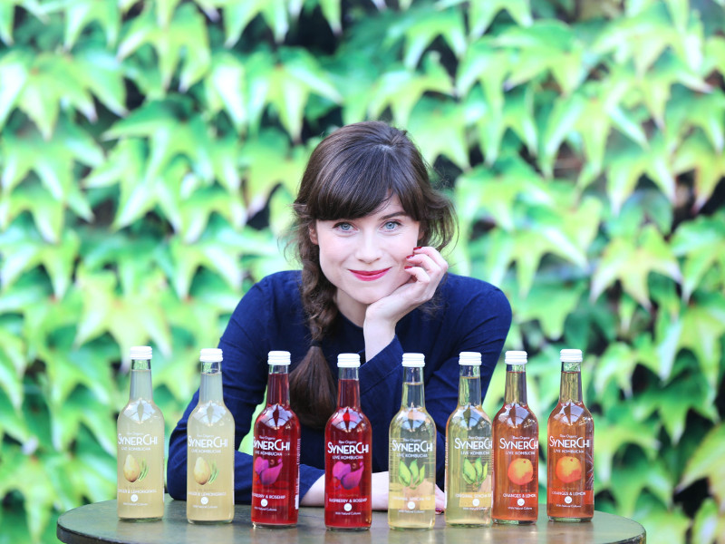 Dark-haired woman behind bottles of Kombucha.