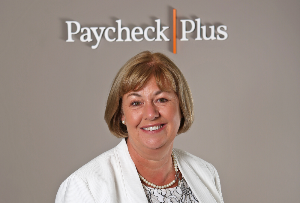 blonde-haired woman in white coat under sign Paycheck Plus.