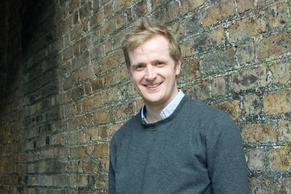 Fair-haired man in green jumper beside a brick wall.