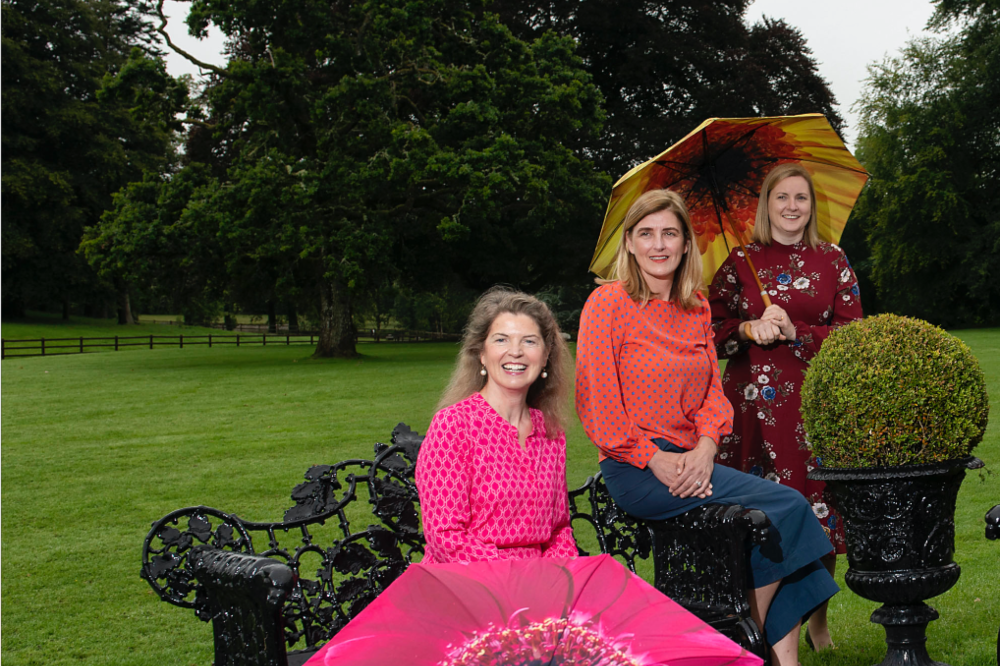 Three women seated on a park bench with colourful pink umbrellas.
