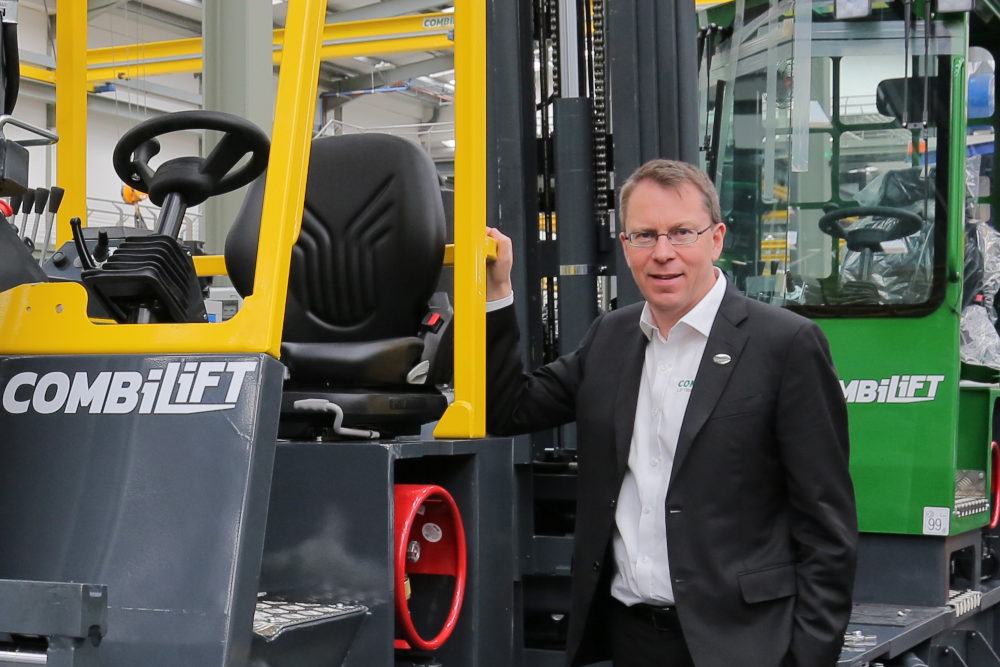 Man in black suit standing beside a forklift.