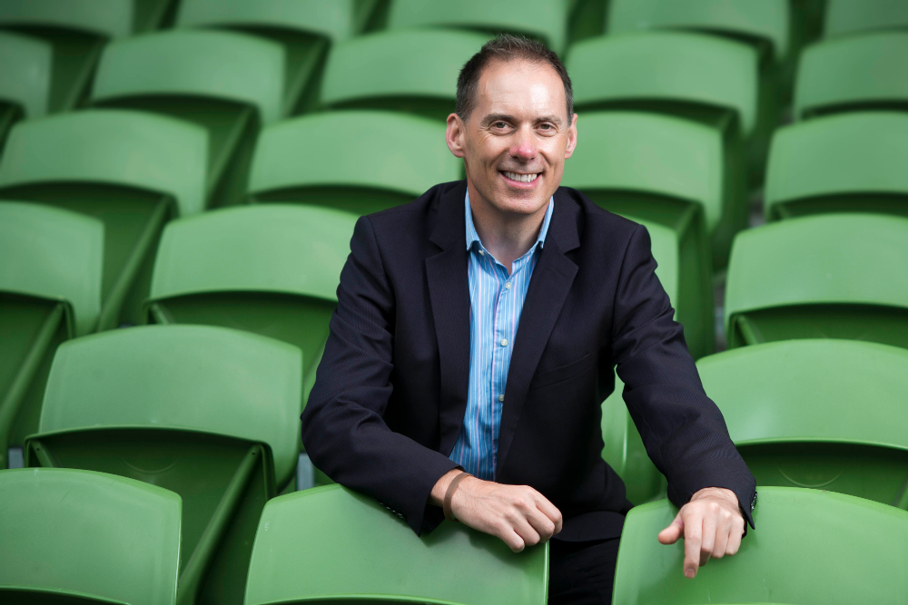 Man in dark suit sitting amid green chairs.