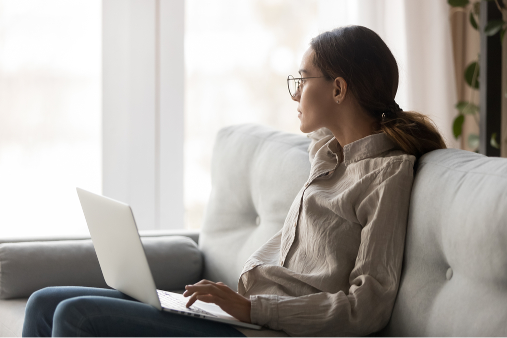 Young woman sitting on couch with laptop looking out window.