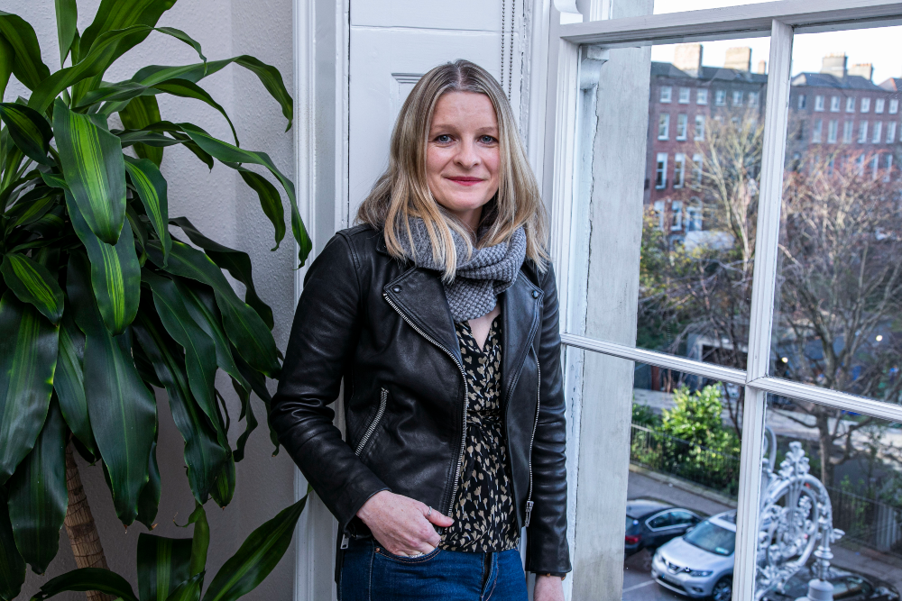 Blonde woman in leather jacket standing beside a window.