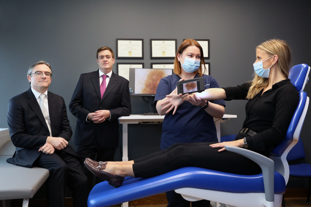 Two men in suits beside a dermatological nurse and her patient.