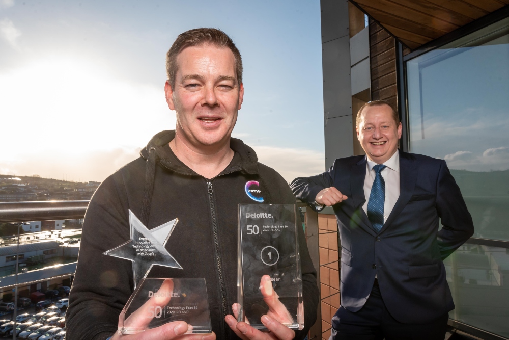 Man in brown hoodie holding two awards beside man in suit,