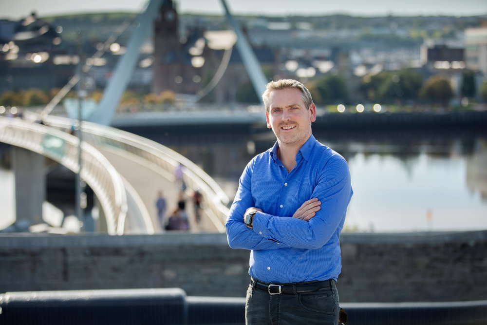 Man in blue shirt beside river and bridge in Derry.