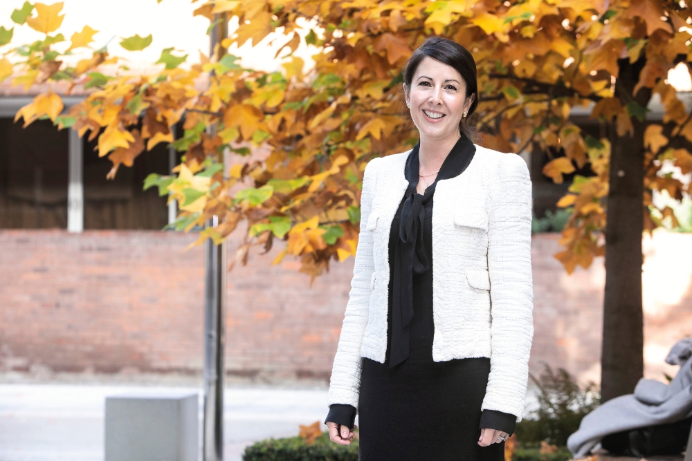Dark-haired woman in white jacket in front of autumnal trees.