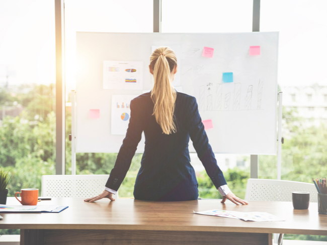 Woman at desk looking at a white board of business objectives.