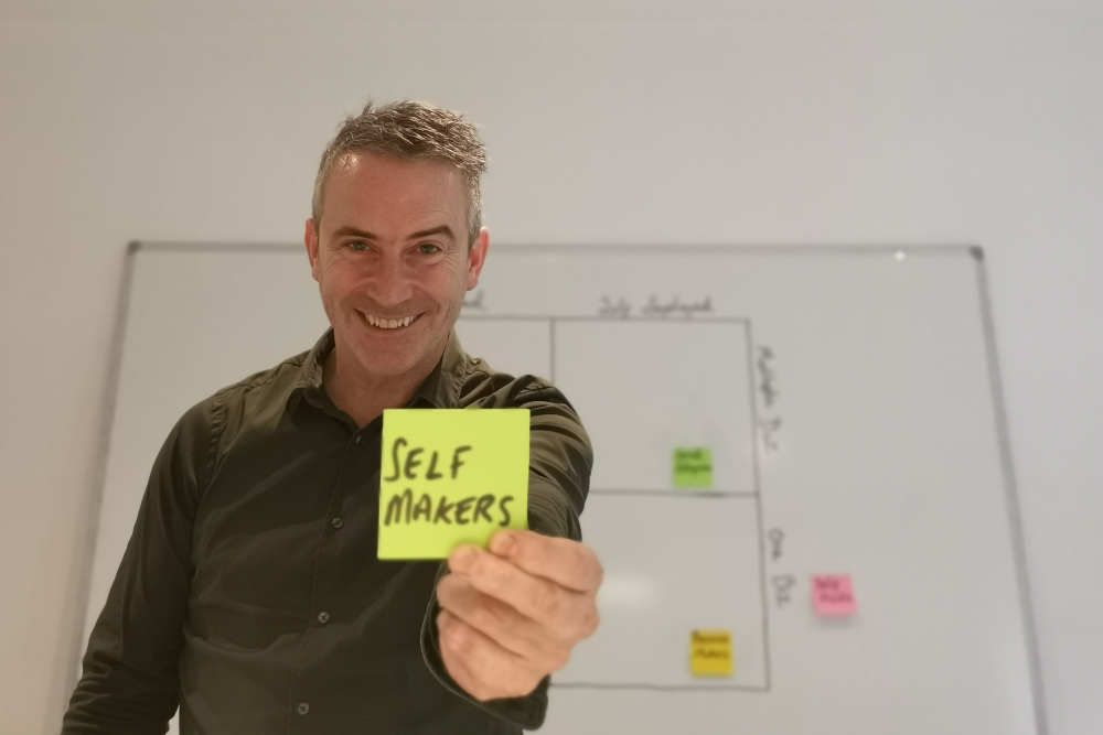 Man in green shirt holding a sticky note saying Self Makers.