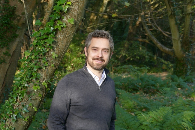 Man in grey jumper standing in woods.