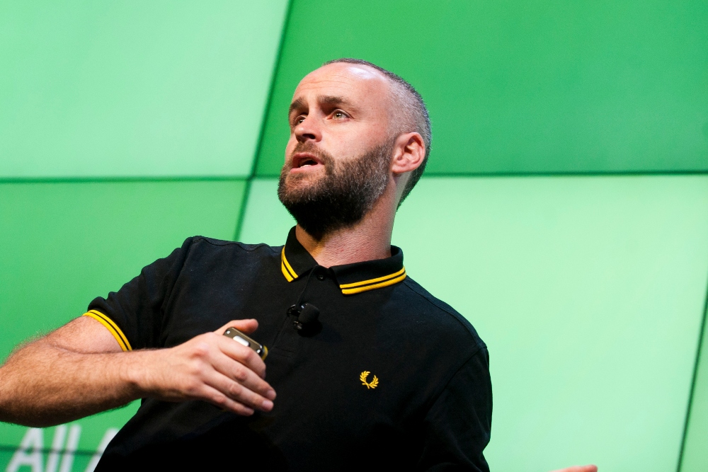 Man in black t-shirt speaking on stage.