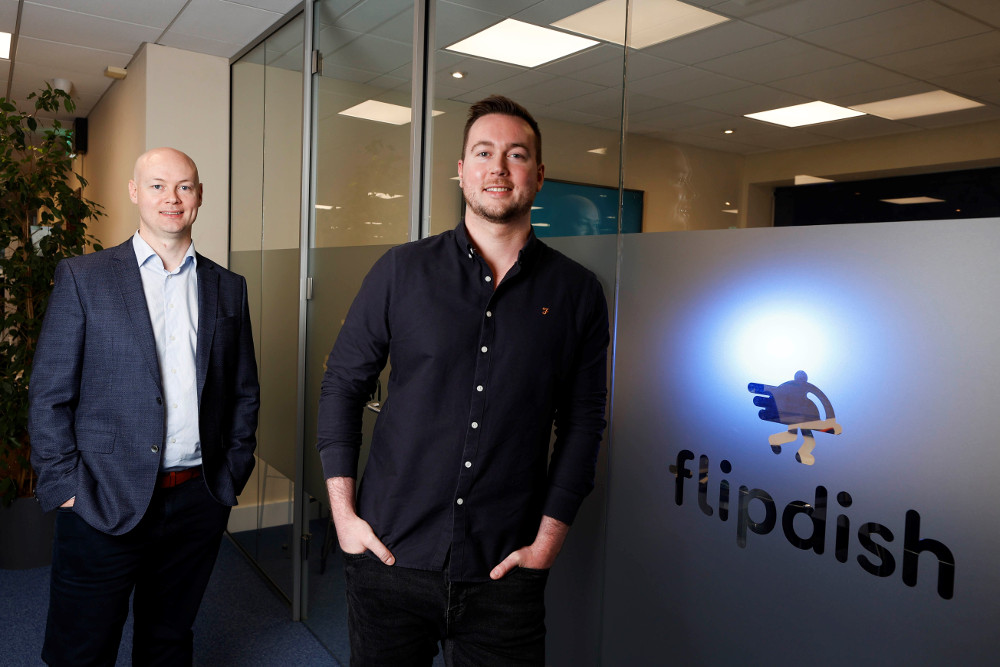 Two men standing beside Flipdish logo on glass window.