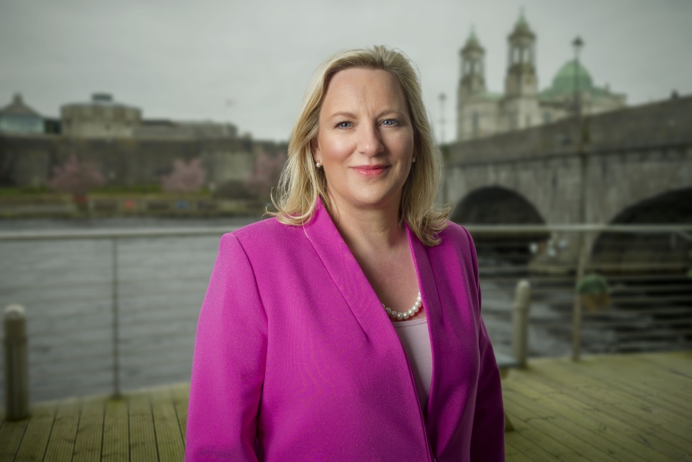 Blonde-haired woman in pink coat standing beside a river.