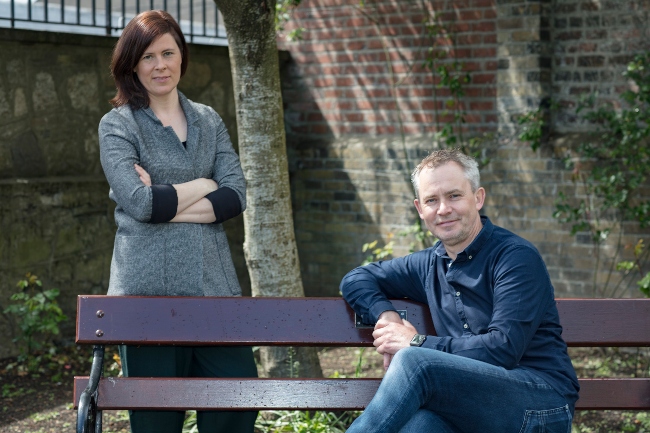 Woman standing behind a bench on which a man is sitting.