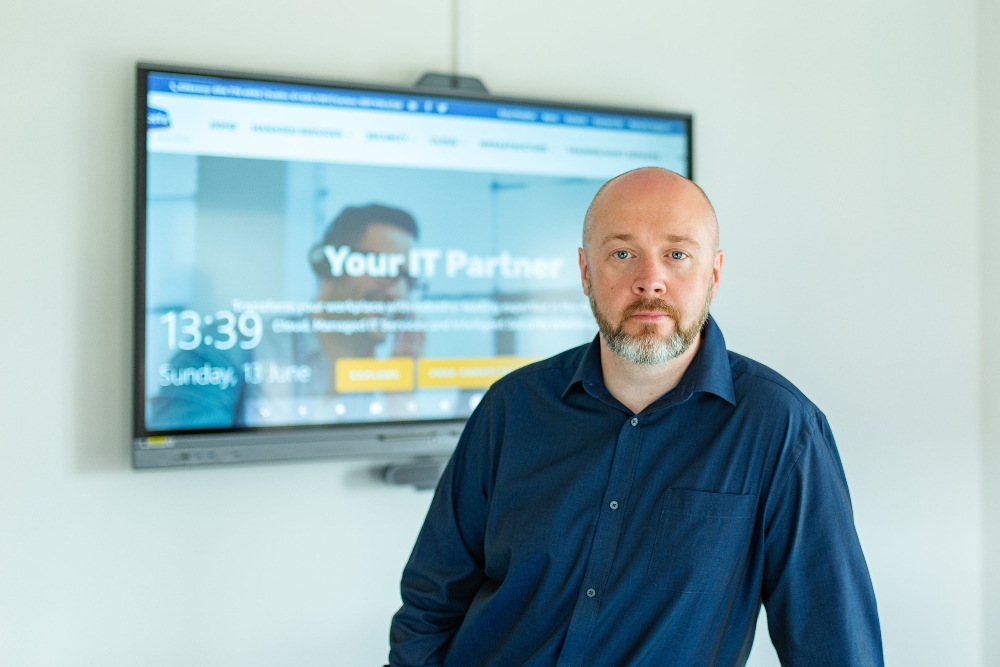 Man in blue shirt standing in front of a computer screen.