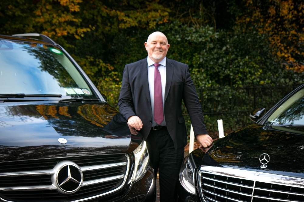 Man in suit standing beside two Mercedes luxury vehicles.