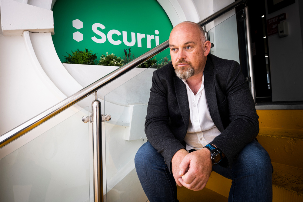 Bearded man sitting on stairs under Scurri logo.