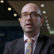 Man in grey suit wearing glasses.