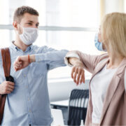 Millennial man and woman in protective masks are touched by elbows in interior of office during COVID-19 epidemic.