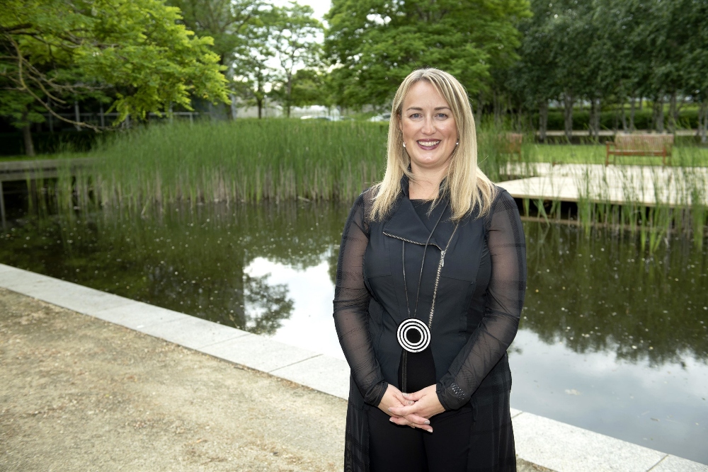Fair haired woman in black dress beside a pond.