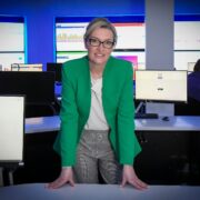 Woman in green jacket with digital screens in background.