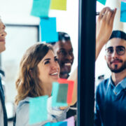 Diverse team of positive young people laughing while working together during brainstorming and standing behind glass wall with sticky colorful papers.
