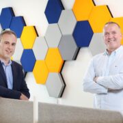 Two men in front of a patterned wall.