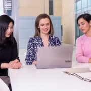 Three women sit around a computer.