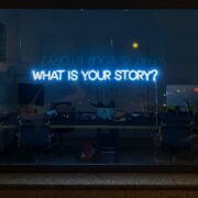 Words 'what's your story' in blue neon on an office window.